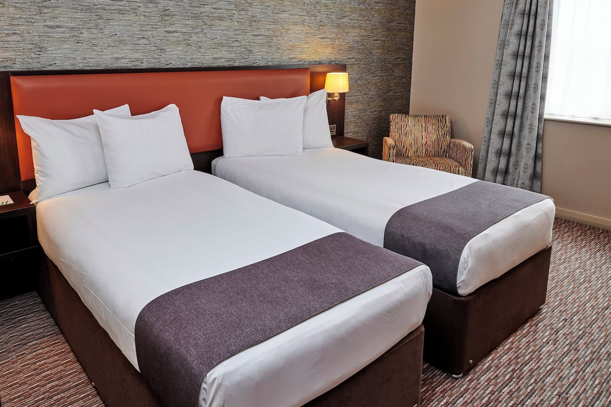 A hotel room with two beds, a nightstand, a lamp, a patterned armchair, and a window with curtains.