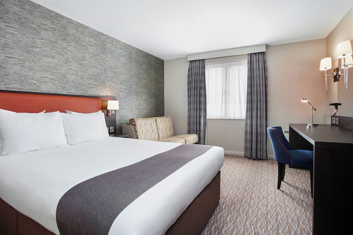 A hotel room with a large bed, a desk, a chair, a lamp, and a window with curtains.