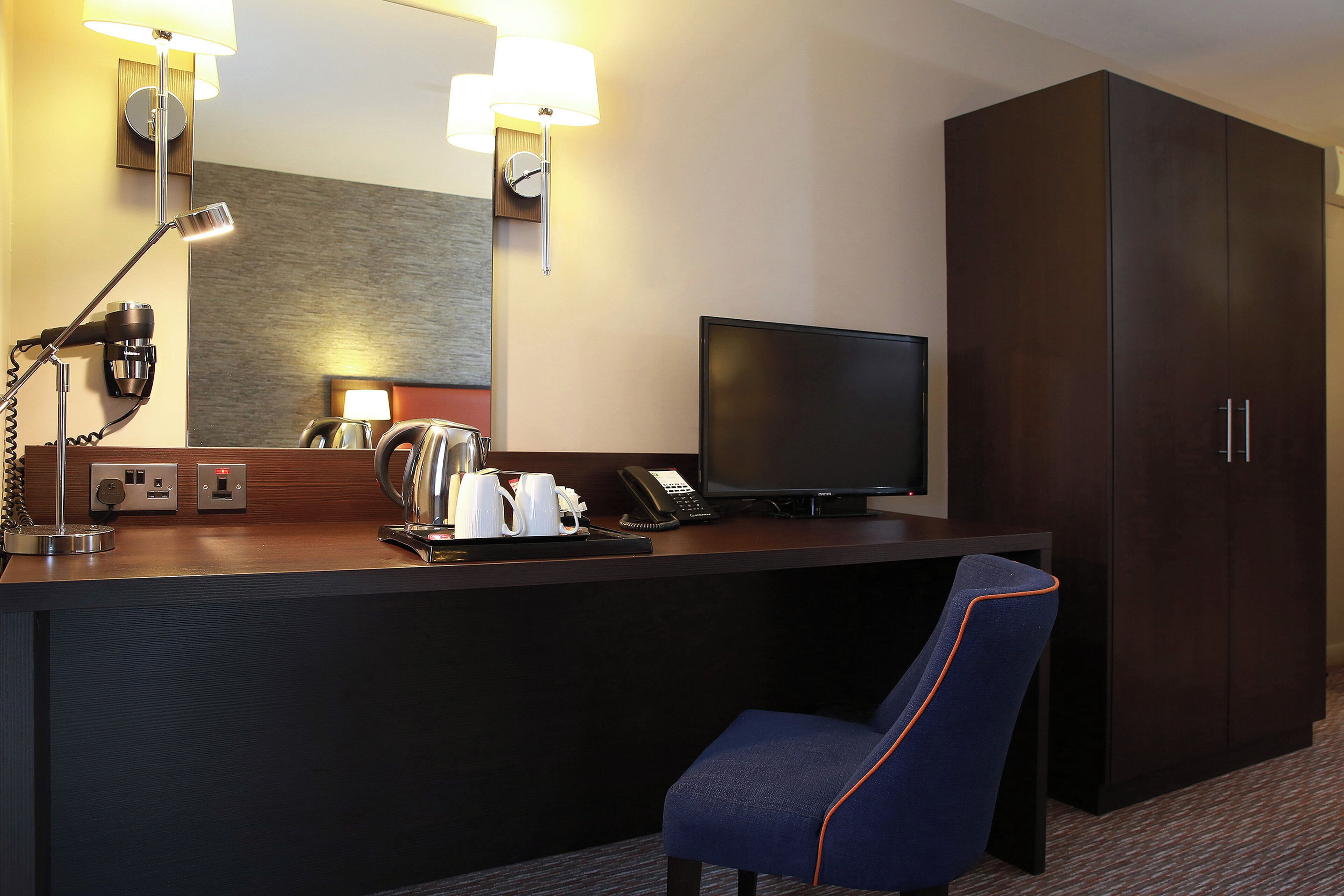 A hotel room with a desk, a television, a chair, a wardrobe, and a mirror.