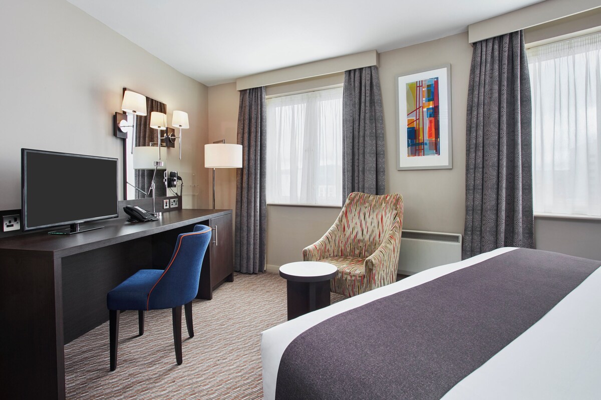 A hotel room with a bed, a desk with a television, a chair, a lamp, and a painting on the wall.