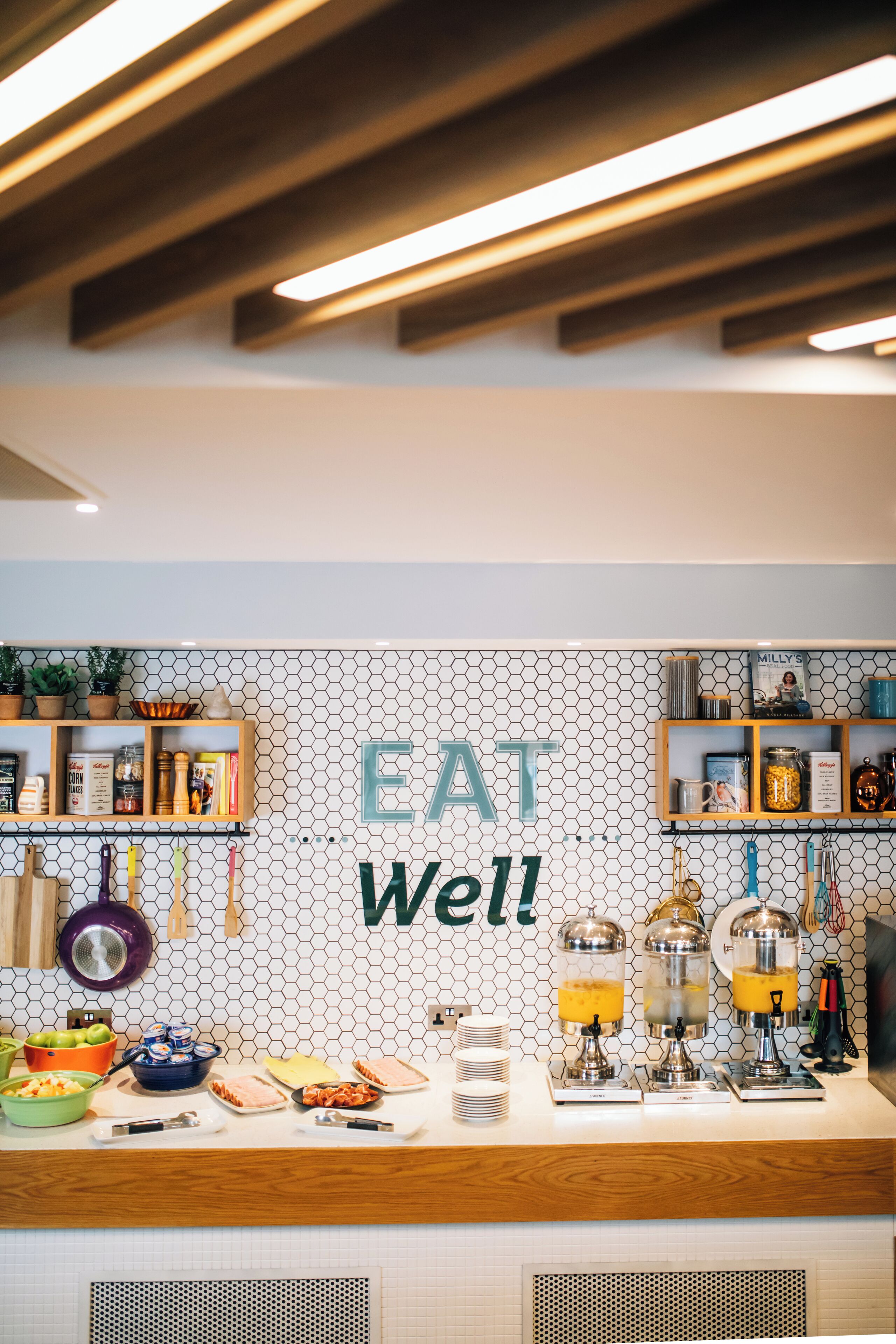 A breakfast bar with a hexagonal tile backsplash, a wooden shelf with kitchen items, and a sign that reads "EAT Well!"