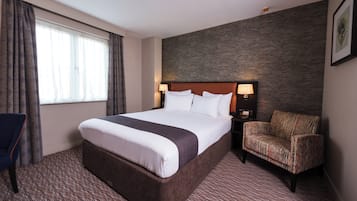 A hotel room with a large bed, a chair, a desk with a lamp, and a window with curtains.