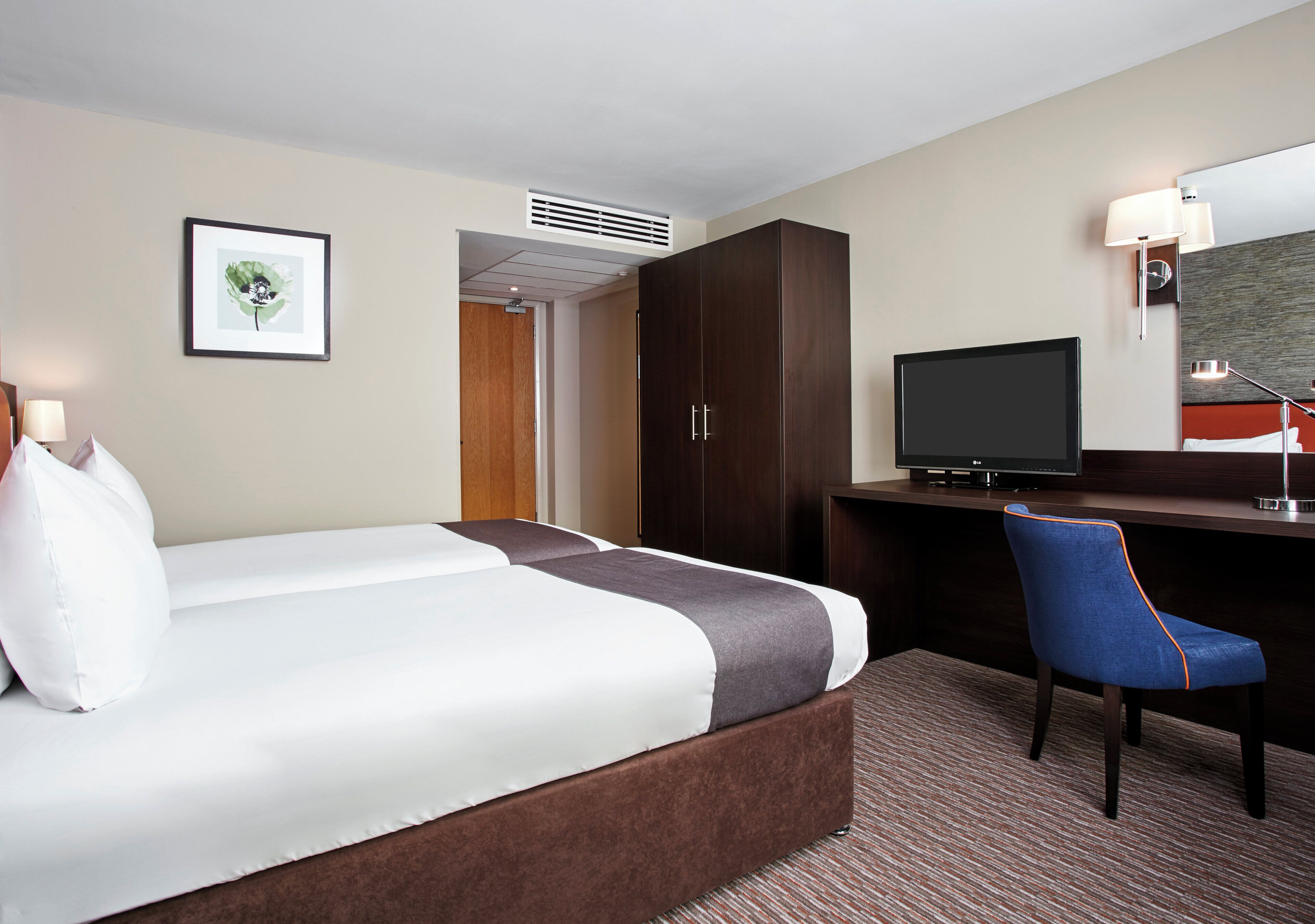 Une chambre d’hôtel avec un lit, un bureau, une chaise, une télévision et un tableau encadré au mur.