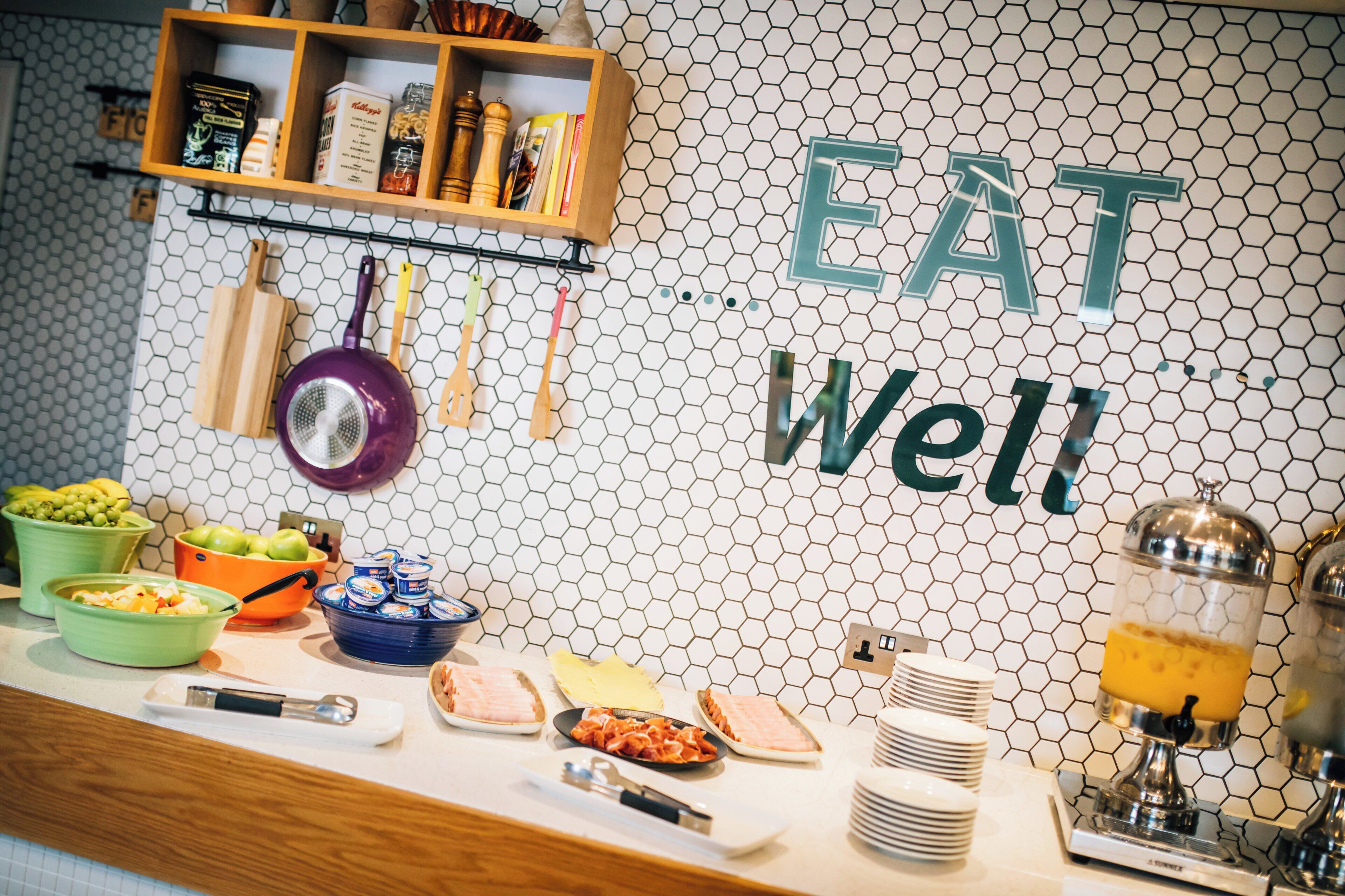 A buffet setup with food items and utensils, a hexagonal tiled wall with the words 'EAT Well' prominently displayed.