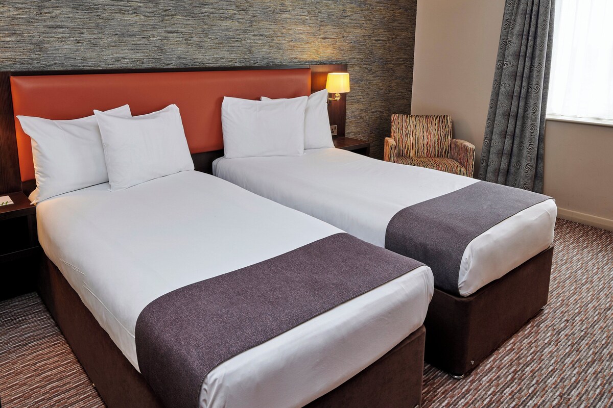 A hotel room with two beds, a nightstand, a lamp, a patterned armchair, and a window with curtains.