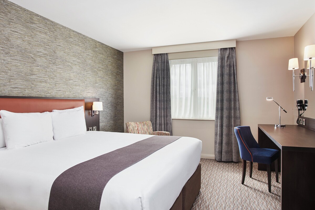 A hotel room with a large bed, a desk, a chair, a lamp, and a window with curtains.