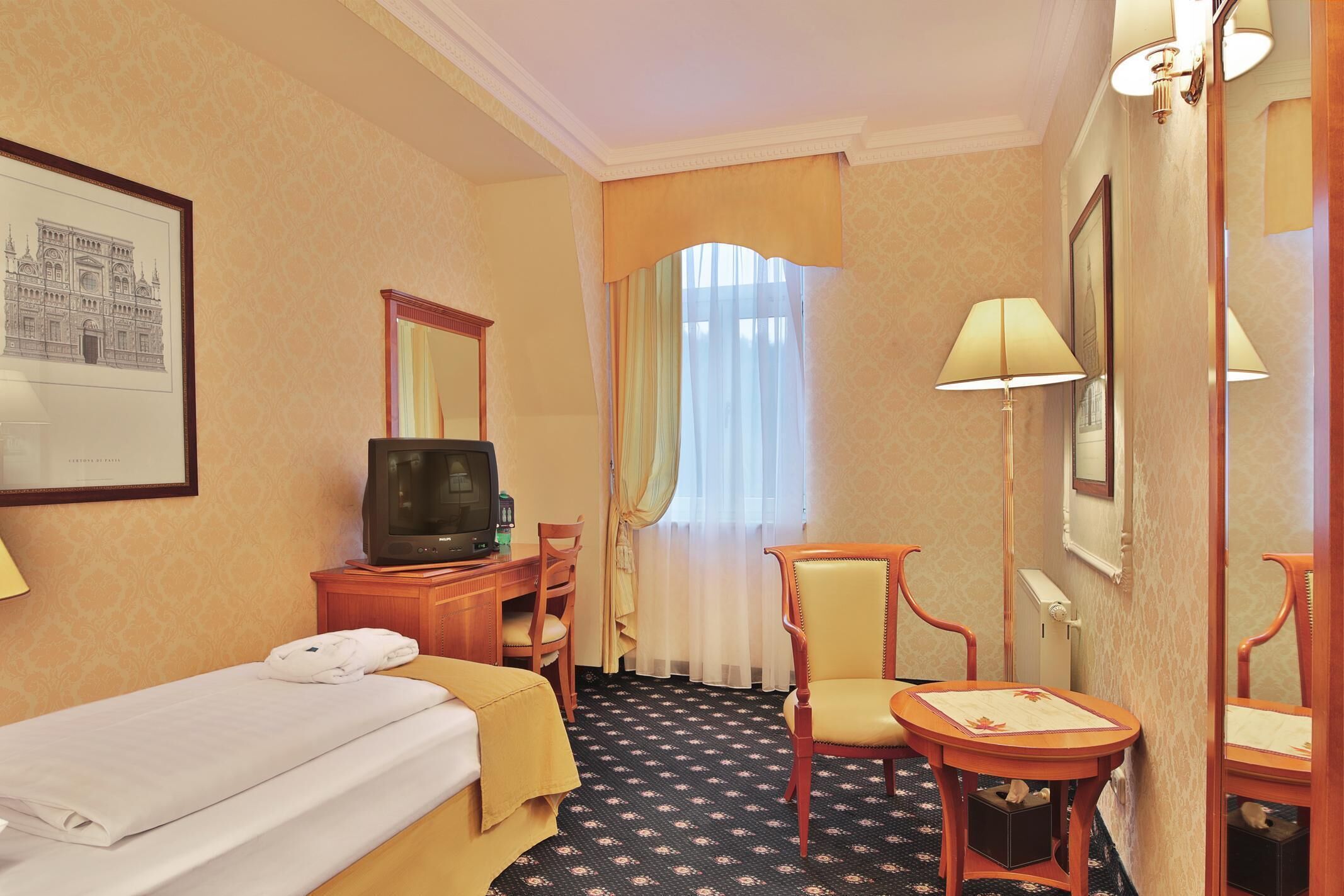 A hotel room with a bed, a desk with a television, a chair, a small table, and a window with curtains.