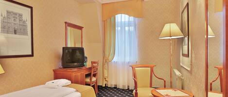 A hotel room with a bed, a desk with a television, a chair, a small table, and a window with curtains.
