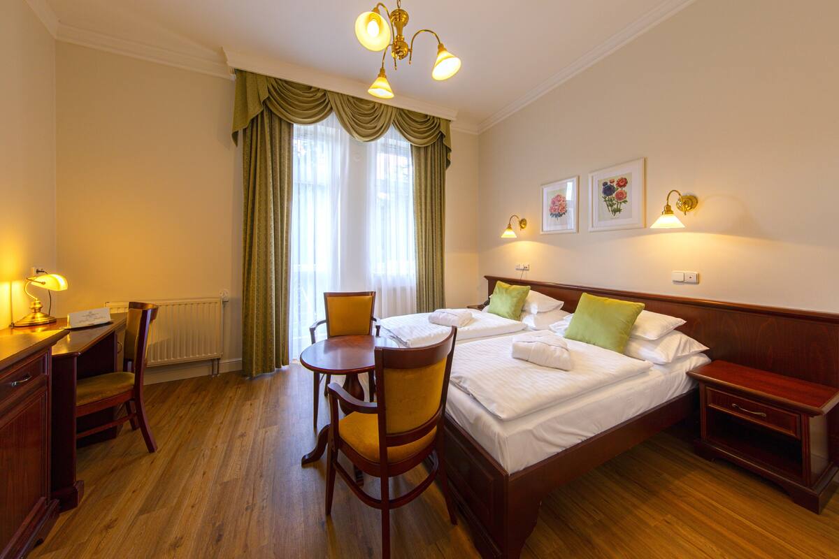 Een hotelkamer met een tweepersoonsbed, twee nachtkastjes, een bureau met stoel, en een raam met gordijnen.