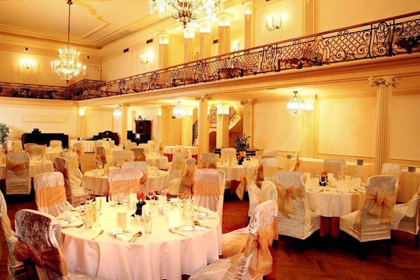 A banquet hall with round tables, chairs with white covers, chandeliers, and a balcony with ornate railings.