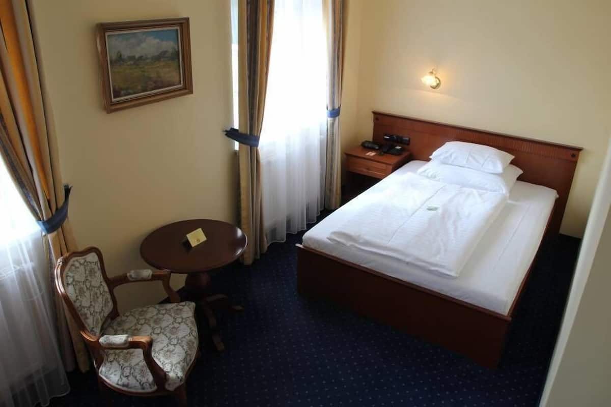 Een hotelkamer met een bed, een stoel, een klein tafeltje, een nachtkastje, een lamp en een schilderij aan de muur.