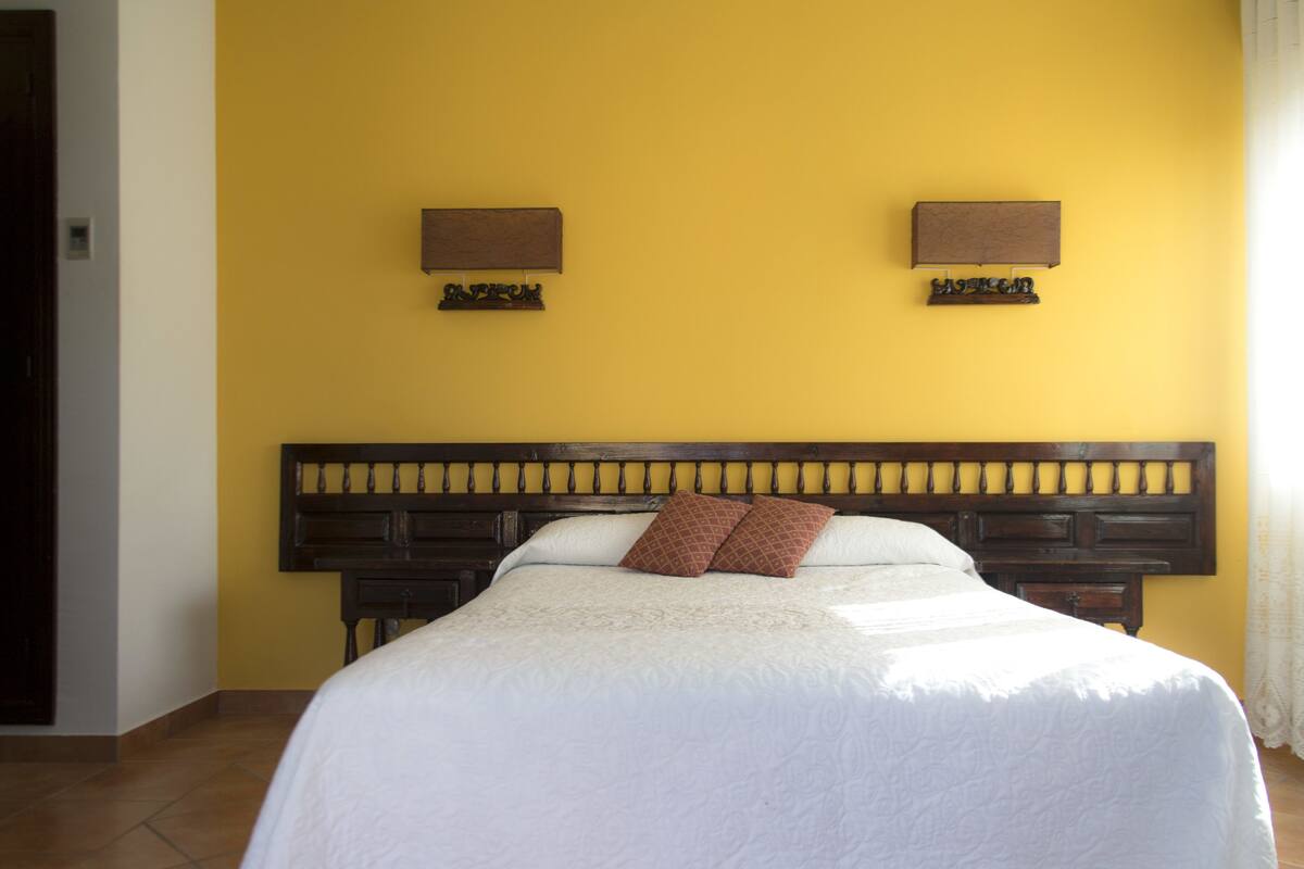 A neatly made bed with a wooden headboard, two wall-mounted lamps, and a plain yellow wall.