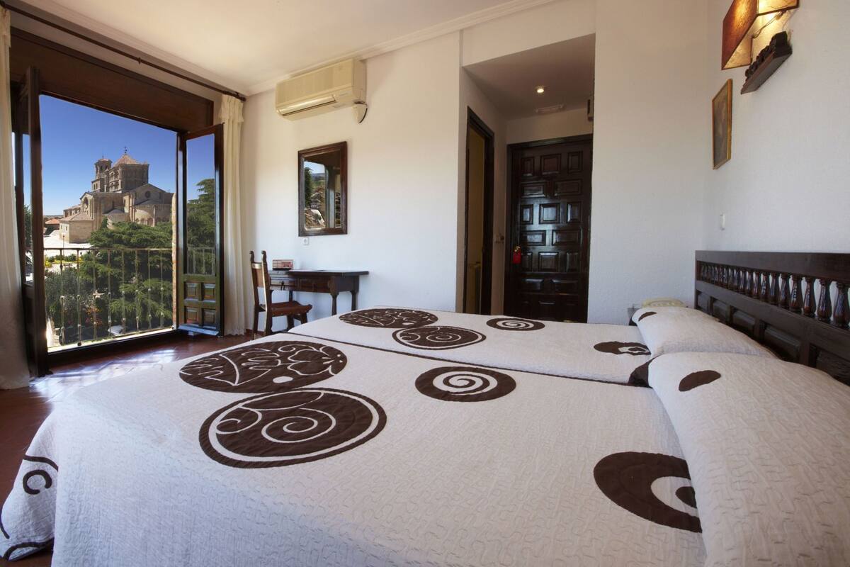A hotel room with a large bed, a desk, a chair, a door, and a view of a building through a balcony.