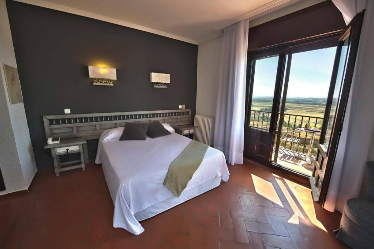 A hotel room with a large bed, a nightstand, a desk, and a view of the countryside through a sliding glass door.