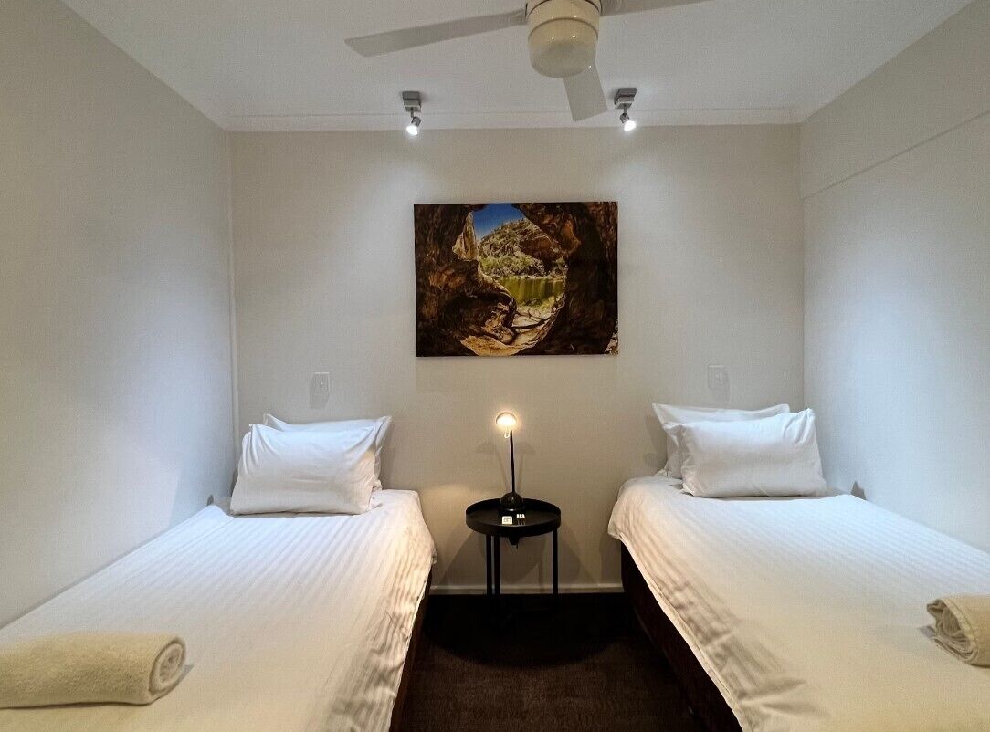 Une chambre d’hôtel avec deux lits simples, une petite table avec une lampe, un ventilateur de plafond et un tableau au mur.
