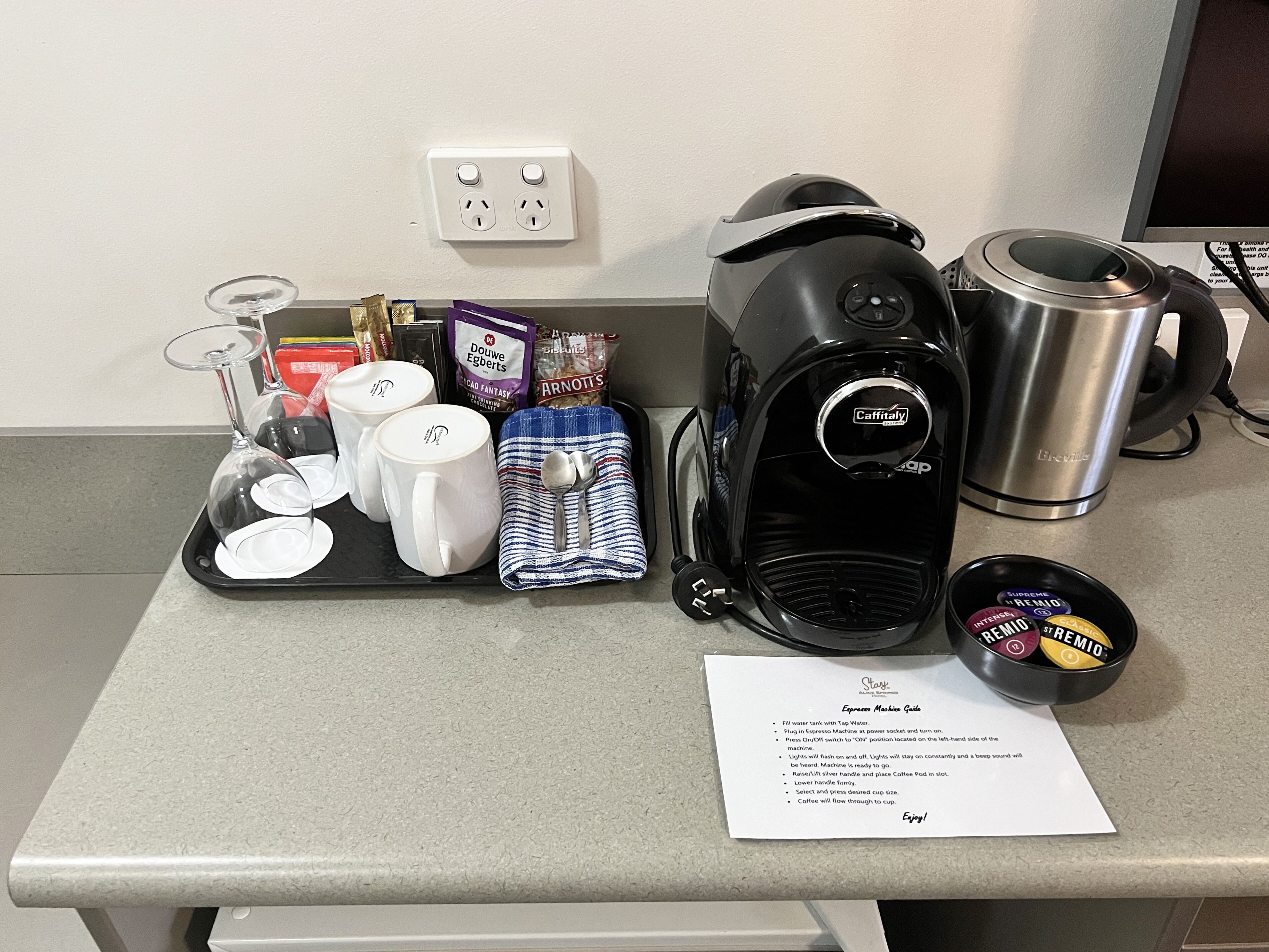 A coffee station with a coffee maker, cups, a spoon, a kettle, and a coffee pod container.