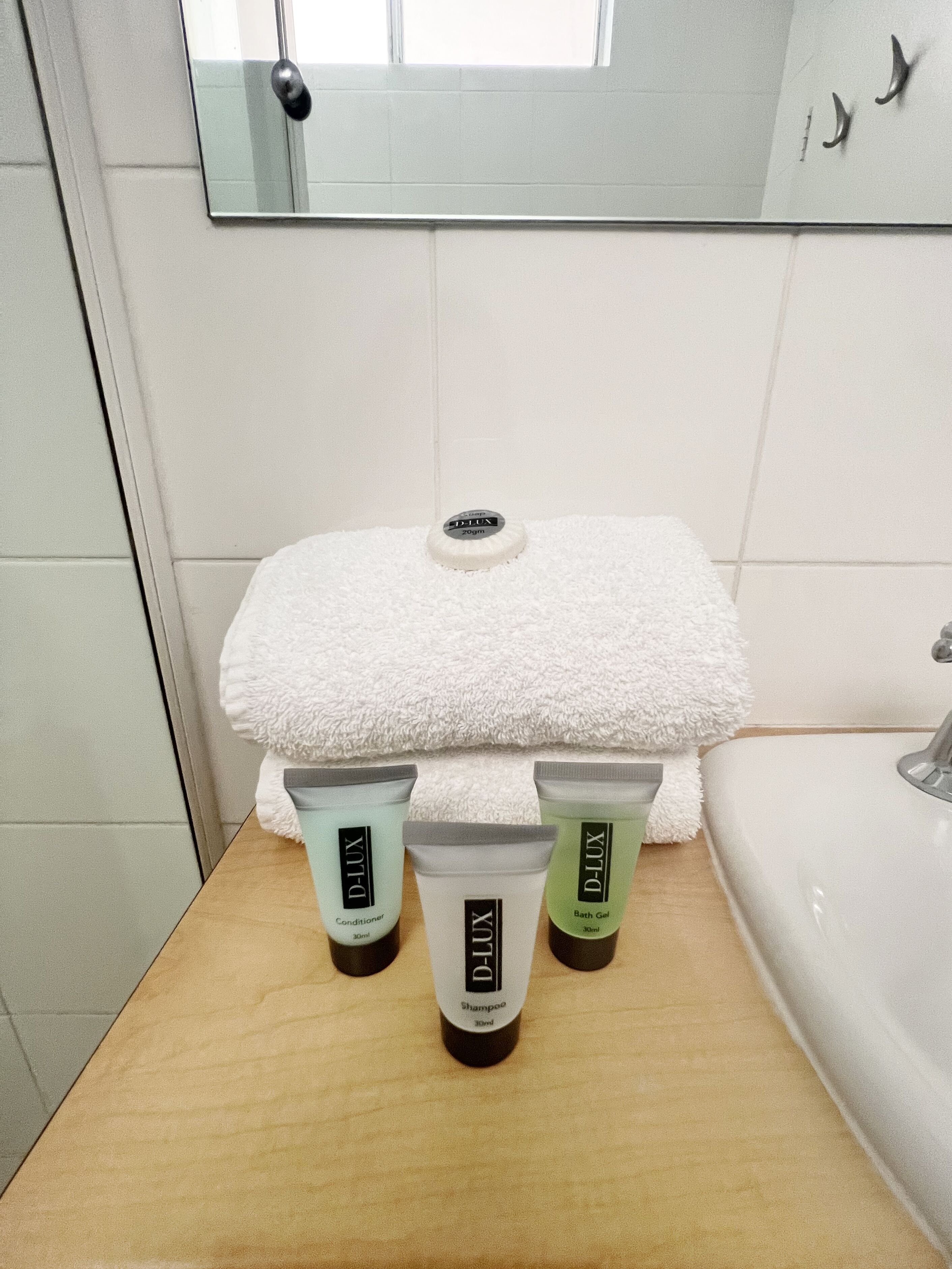 A bathroom with a white towel, a mirror, and three D-Lux toiletries on a wooden tray.