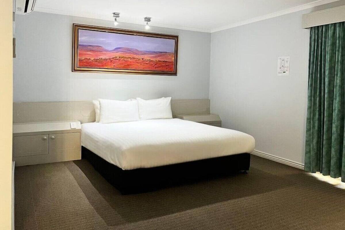 A hotel room with a bed, a nightstand, a wall-mounted painting, and a green curtain.