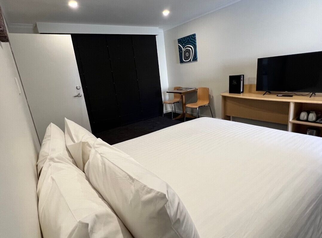 Hotellihuone, jossa on suuri sänky, työpöytä, jossa on tietokone ja televisio, tuoli sekä ovi, joka johtaa pimeään huoneeseen.