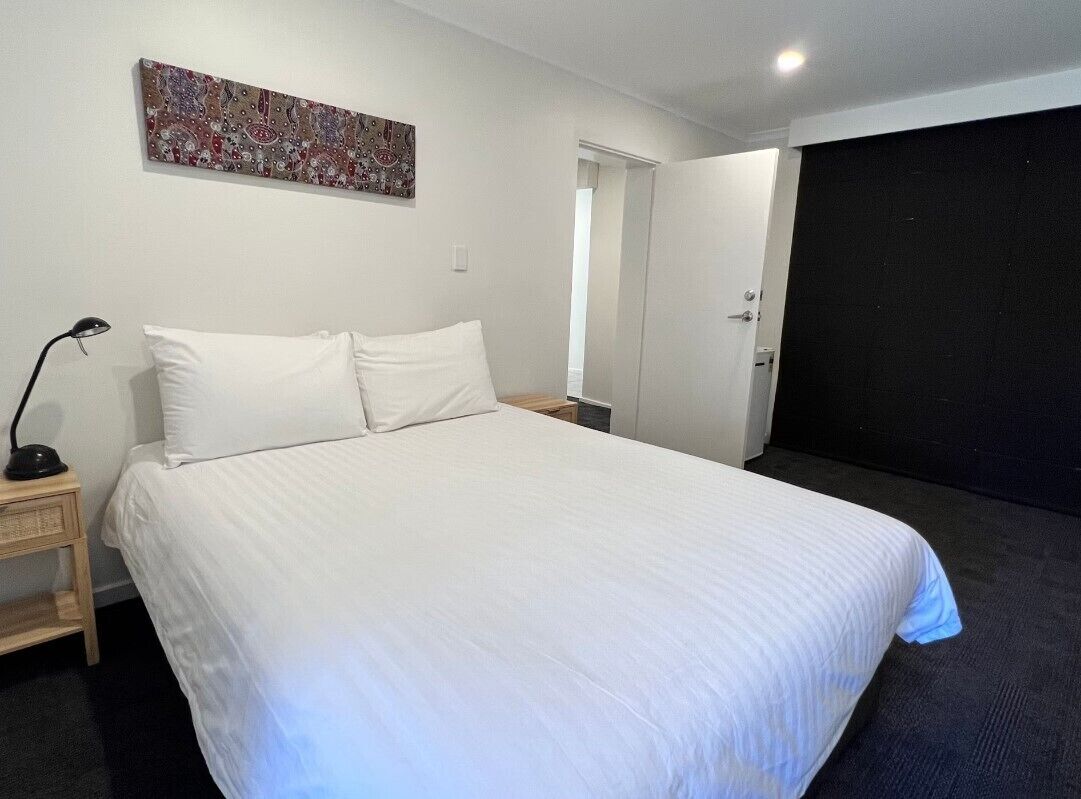 A neatly made bed with white linens in a room with a dark carpet, a small bedside table with a lamp, and a door leading to another room.
