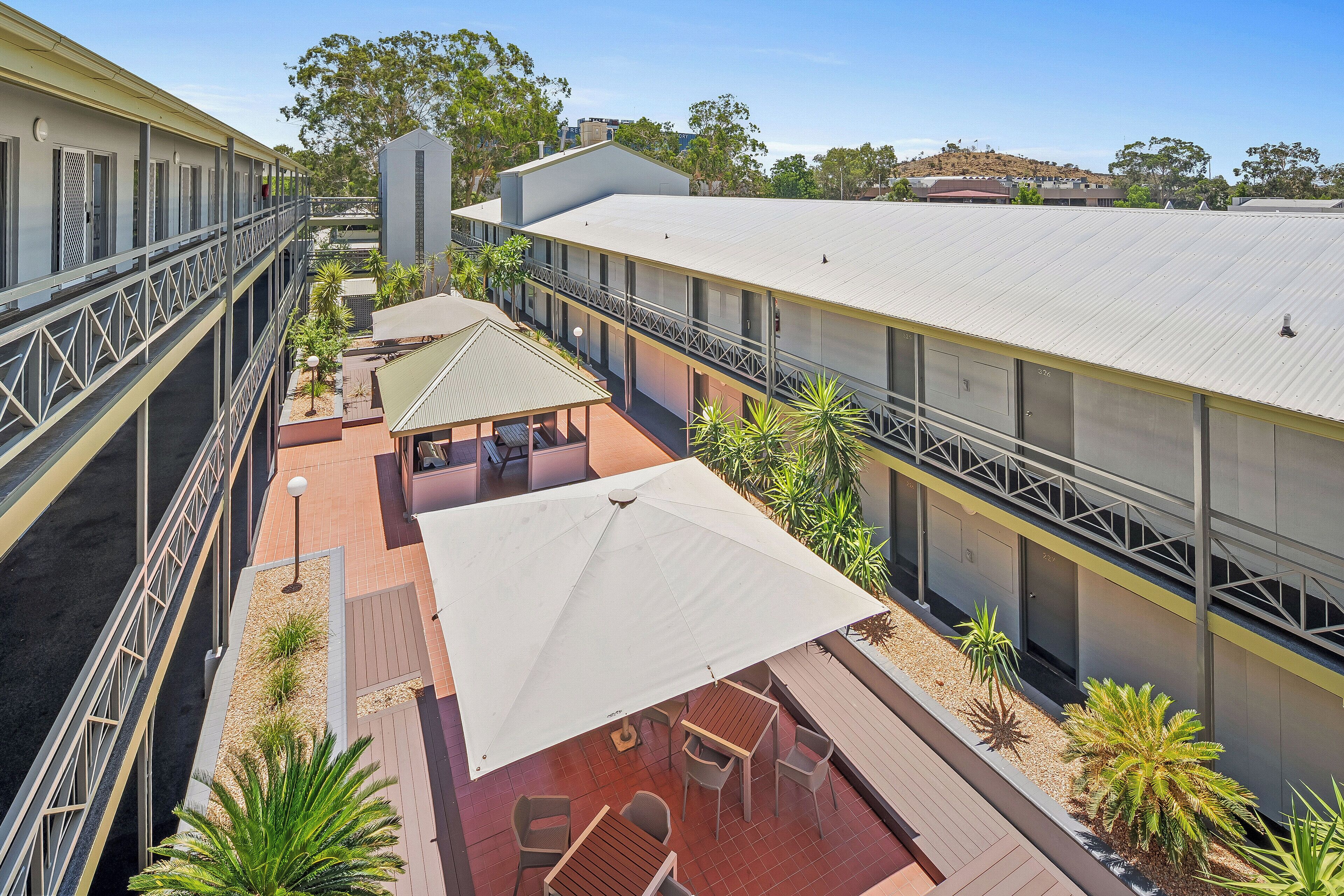 A multi-level outdoor patio area with seating and umbrellas, surrounded by buildings with a flat roof and a balcony.