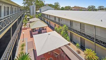 A multi-level outdoor patio area with seating and umbrellas, surrounded by buildings with a flat roof and a balcony.