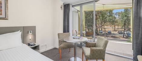 Habitación de hotel con cama, una mesa redonda con dos sillas, una mesita con teléfono y un ventanal con vistas a árboles y coches aparcados.