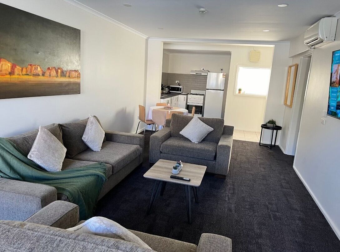 A modern living room with grey sofas, a coffee table, and a painting on the wall. A kitchen is visible in the background.