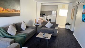 A modern living room with grey sofas, a coffee table, and a painting on the wall. A kitchen is visible in the background.