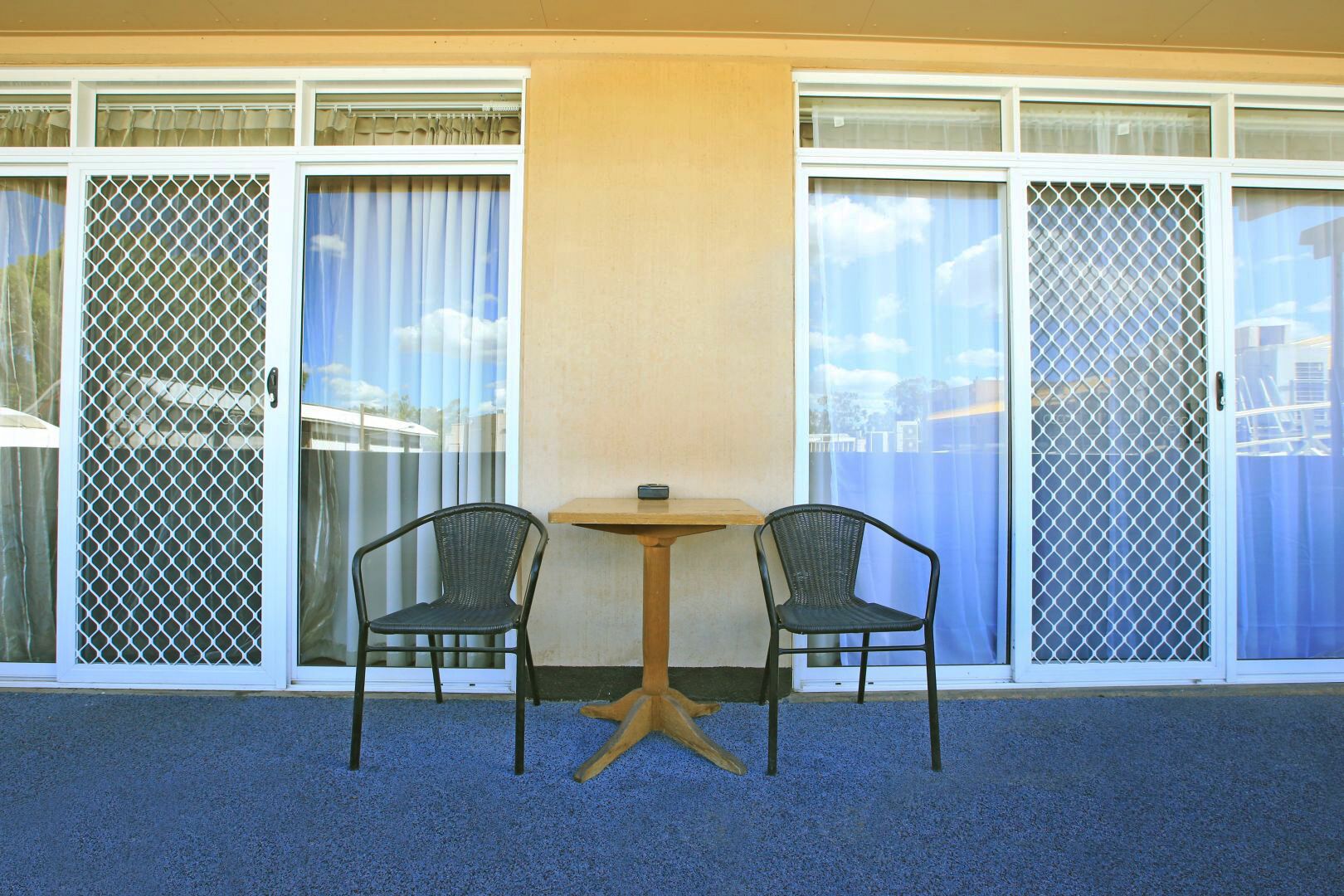 A room with a small table and two chairs, large windows with curtains, and a blue carpet.