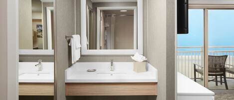 A hotel bathroom with two sinks, a mirror, and a shower. There is a balcony with a chair and a TV.