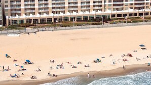 A large hotel with multiple floors and balconies, situated next to a sandy beach with people and umbrellas.