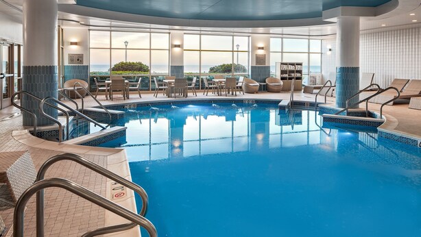 An indoor swimming pool with clear views of the ocean, surrounded by lounge chairs and tables.