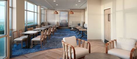 A spacious hotel lobby with multiple seating areas, including round tables and chairs, a large central table, and a view of the outdoors through large windows.