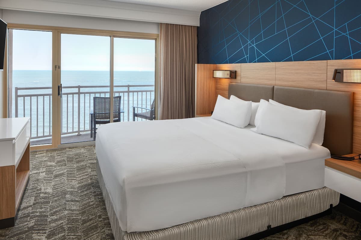 A hotel room with a large bed, a desk, and a chair. The room has a view of the ocean through the sliding glass doors.