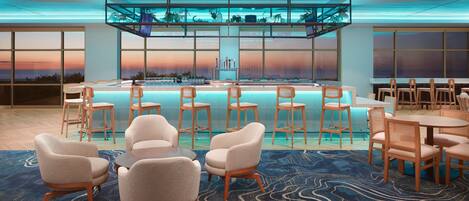 A modern hotel lobby with a pool area, seating arrangements, and large windows offering a view of the sunset.