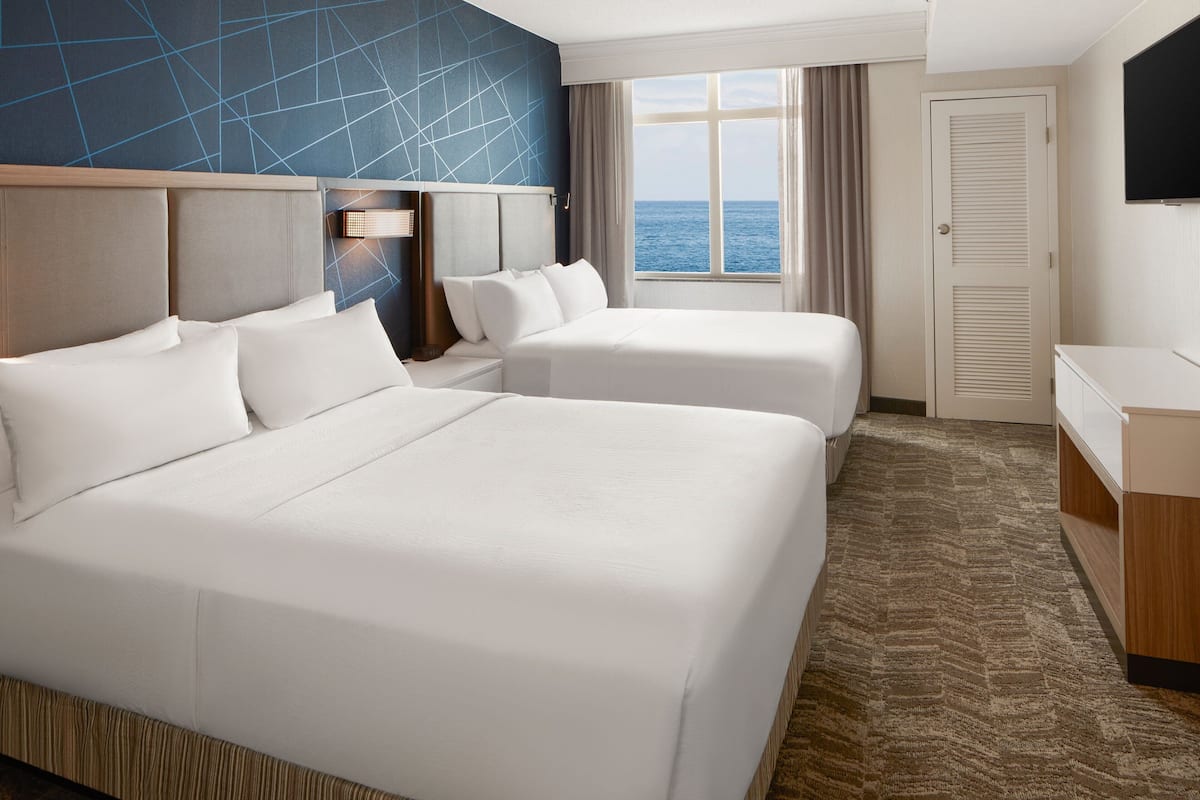 A hotel room with two beds, a TV, a desk, and a view of the ocean.