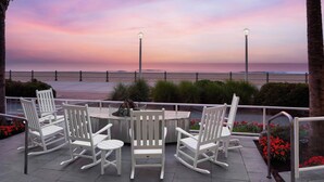A patio with white rocking chairs and a table, overlooking a beach with a sunset.