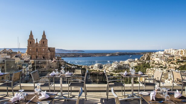 A rooftop restaurant with tables set for dining, overlooking a coastal town and a prominent church.