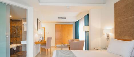 Quarto de hotel com uma cama grande, uma escrivaninha com cadeira, casa de banho com chuveiro e lavatório, e guarda-roupa de madeira.