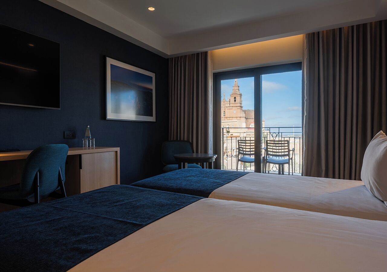 Ein Hotelzimmer mit zwei Betten, einem Schreibtisch, Stühlen und einem Balkon mit Blick auf ein Gebäude.