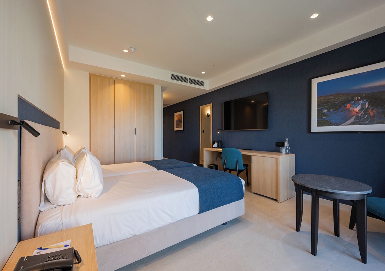 A hotel room with two beds, a desk, a chair, a small table, and a painting on the wall.