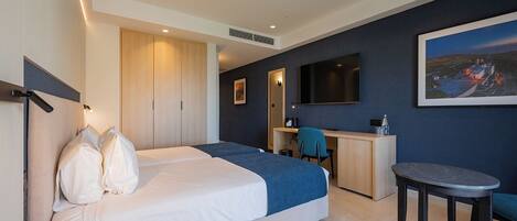A hotel room with two beds, a desk, a chair, a small table, and a painting on the wall.