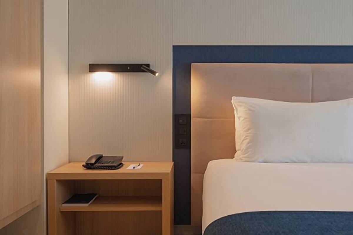 Habitación de hotel con cama, mesita de noche con teléfono y un libro, y una lámpara fijada a la pared.