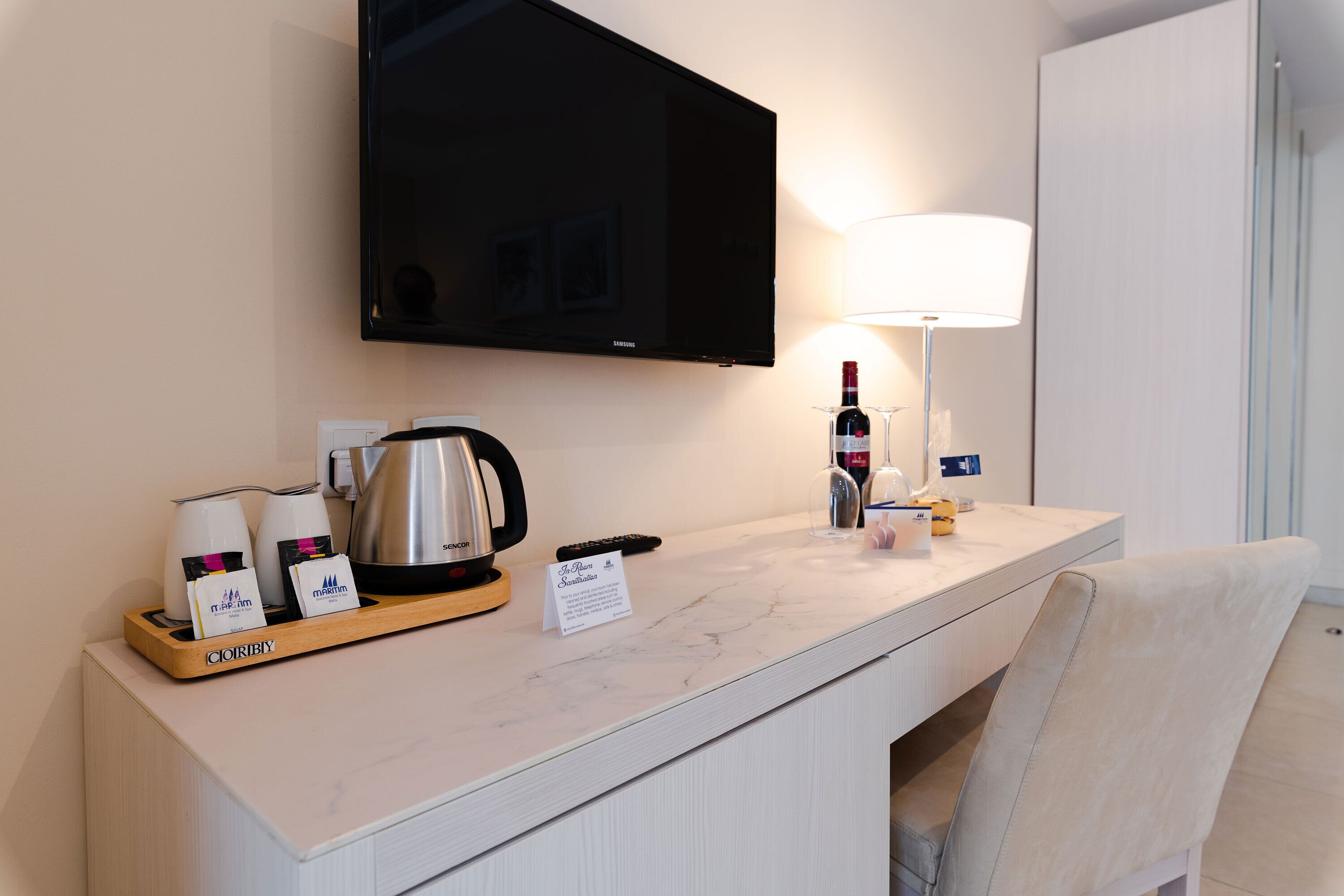 A hotel room with a marble countertop, a flat-screen TV, a coffee maker, a wine bottle, and a lamp.