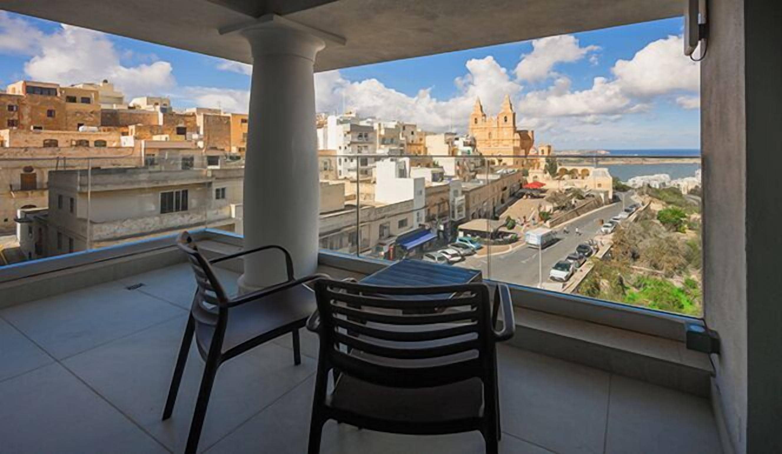 Un balcón con dos sillas y una mesa, con vistas a un paisaje urbano que incluye edificios y una masa de agua.