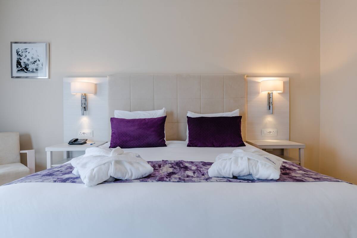 Una habitación de hotel con una cama grande, dos almohadas moradas, toallas blancas y dos mesitas de noche con lámparas.