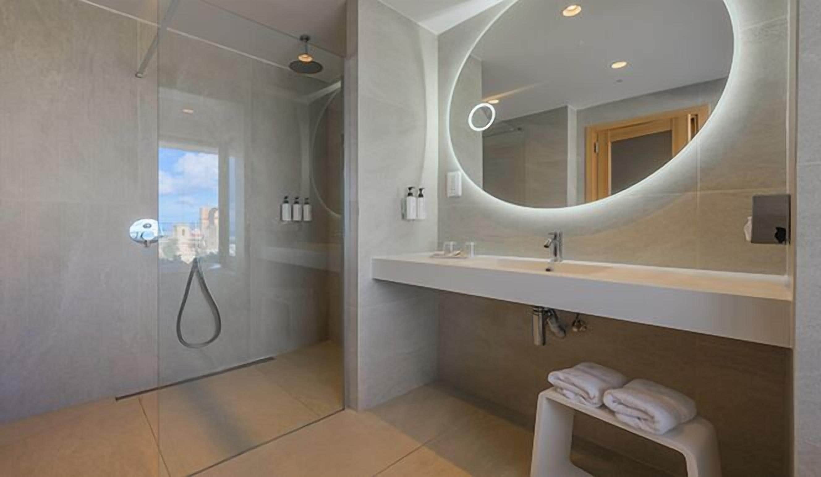A modern bathroom with a glass shower enclosure, a large mirror with a circular light, and a white vanity with a towel rack.