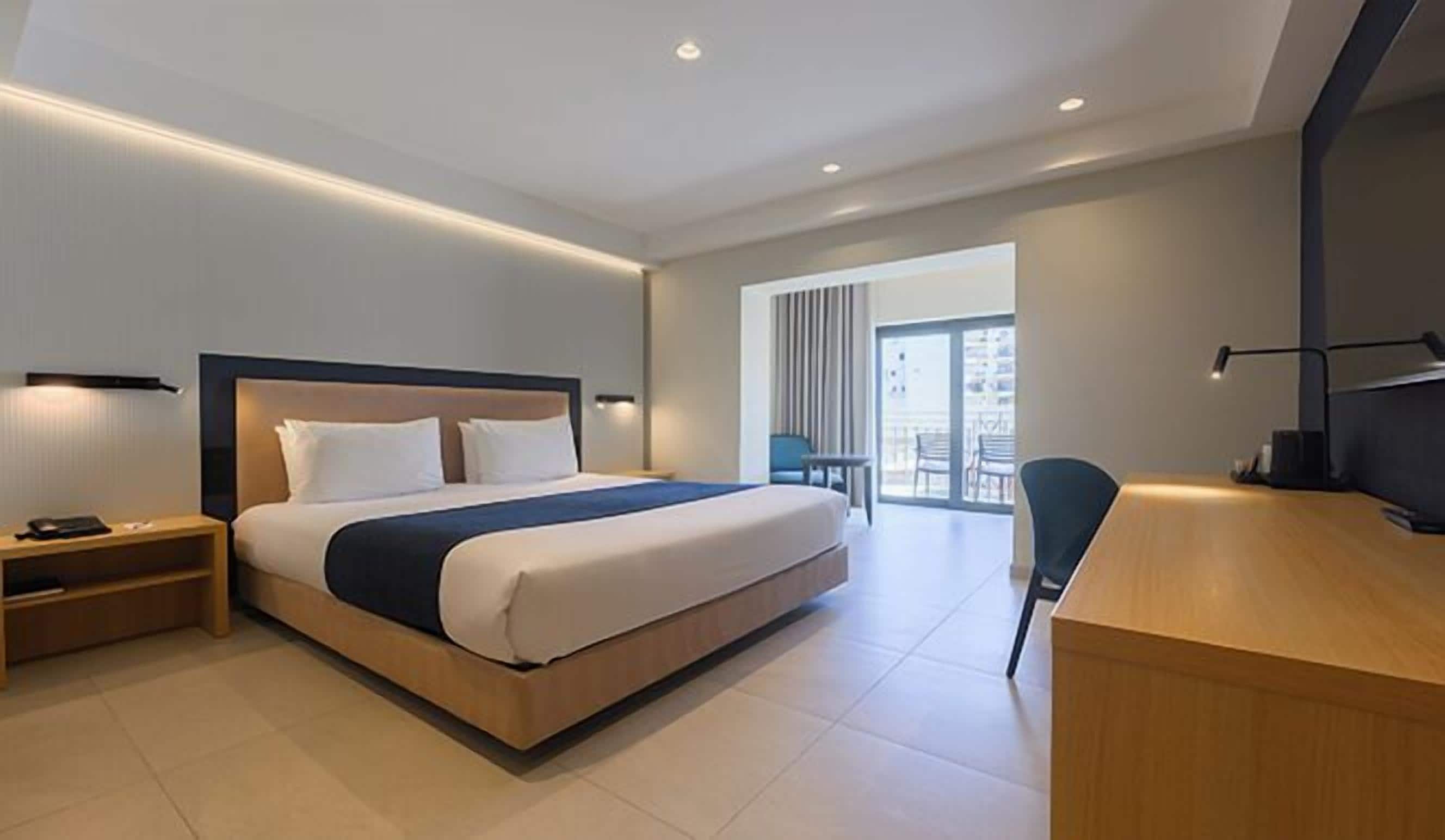 Una habitación de hotel moderna con una cama grande, mesitas de noche, un escritorio y vistas al balcón exterior.