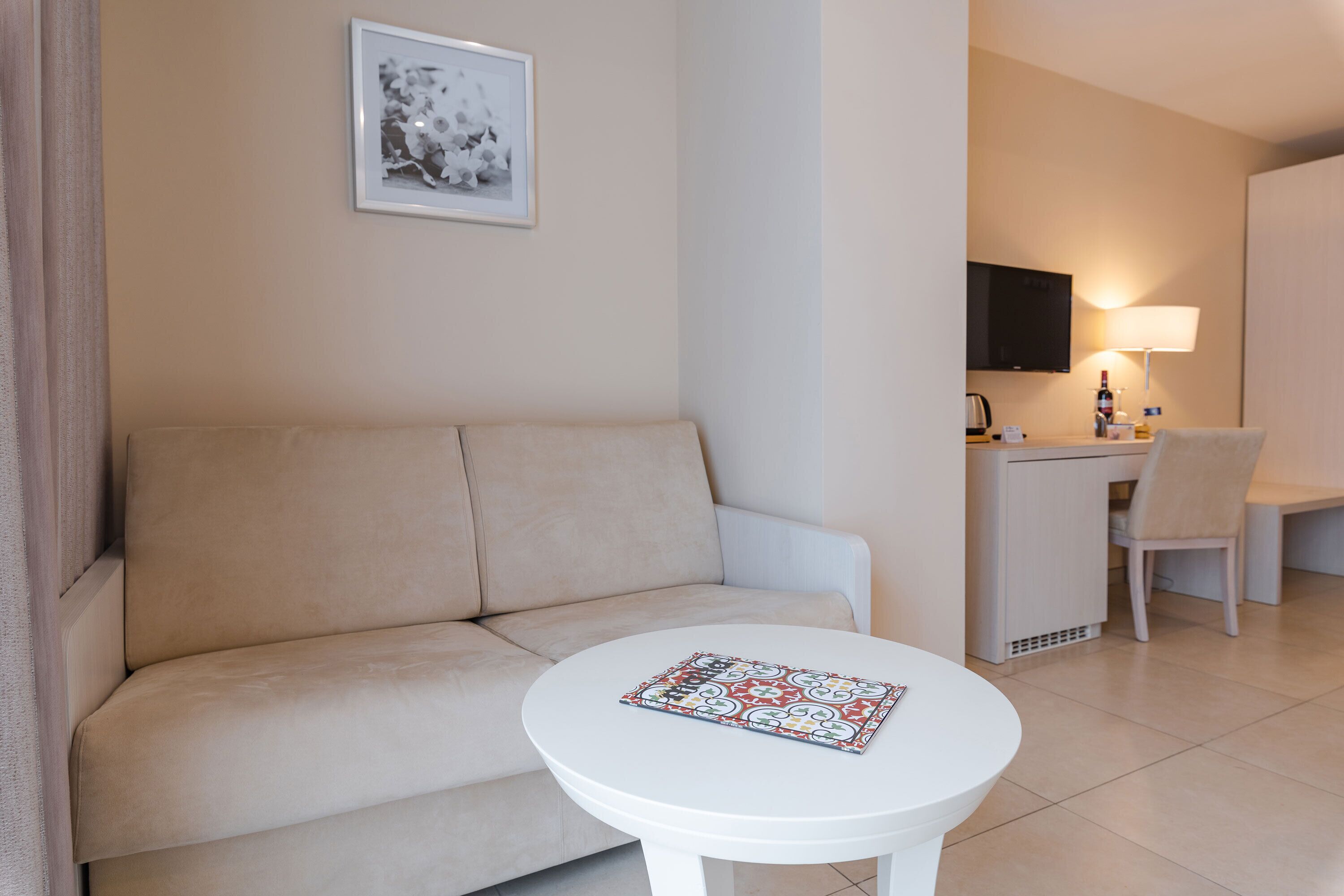A modern hotel room with a beige sofa, a small white table, a desk with a chair, a TV, and a lamp.